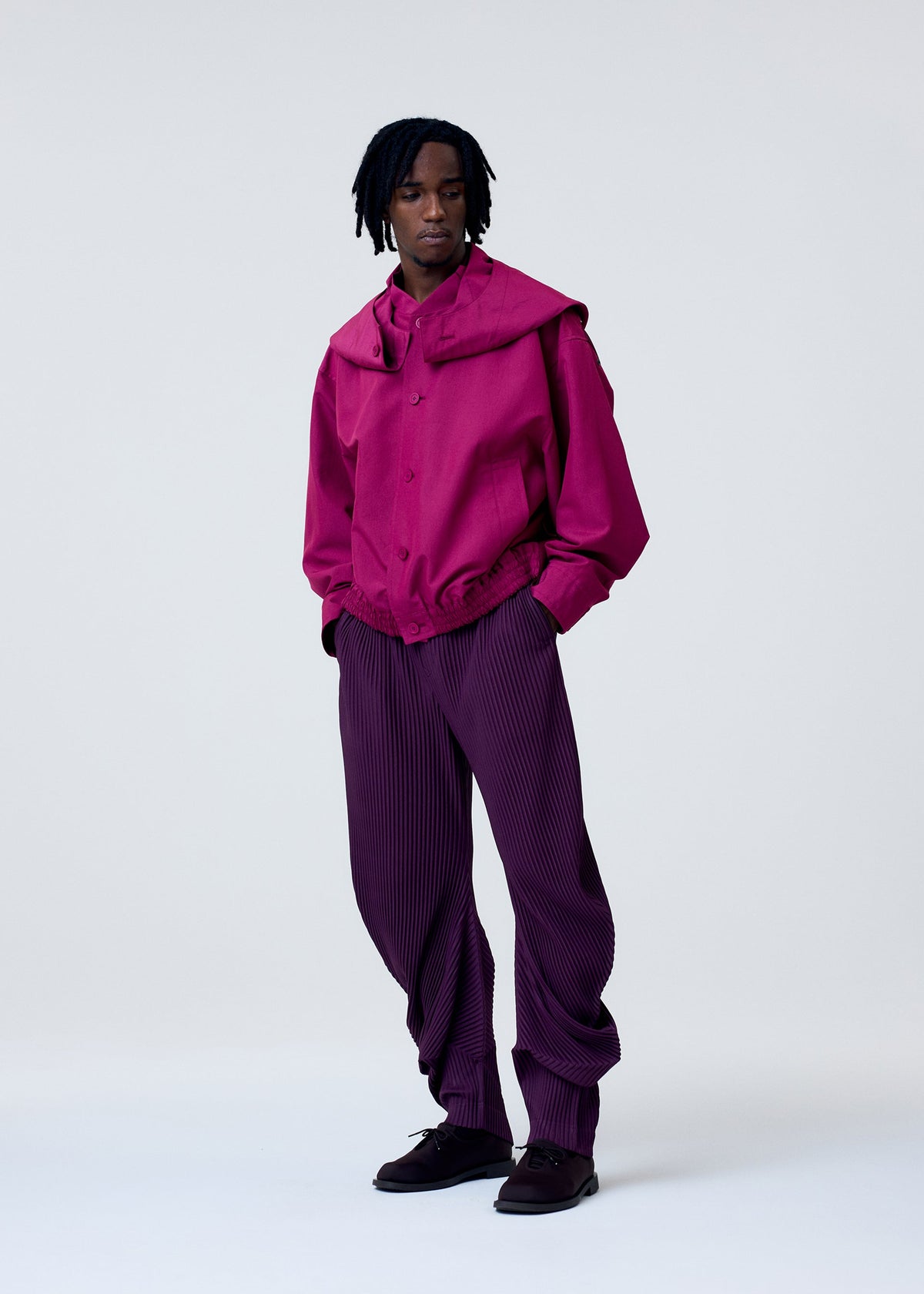 A person with shoulder-length dreadlocks and a neutral expression stands against a light background wearing the CARRIER CARRIED water-repellent jacket and loose pleated trousers by HOMME PLISSÉ ISSEY MIYAKE, paired with black shoes. view 7 of 10