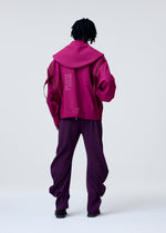 A person with medium-length dreadlocks wears the magenta CARRIER CARRIED hooded jacket and loose, dark purple pleated pants by HOMME PLISSÉ ISSEY MIYAKE, standing with their back to the camera against a plain light background.