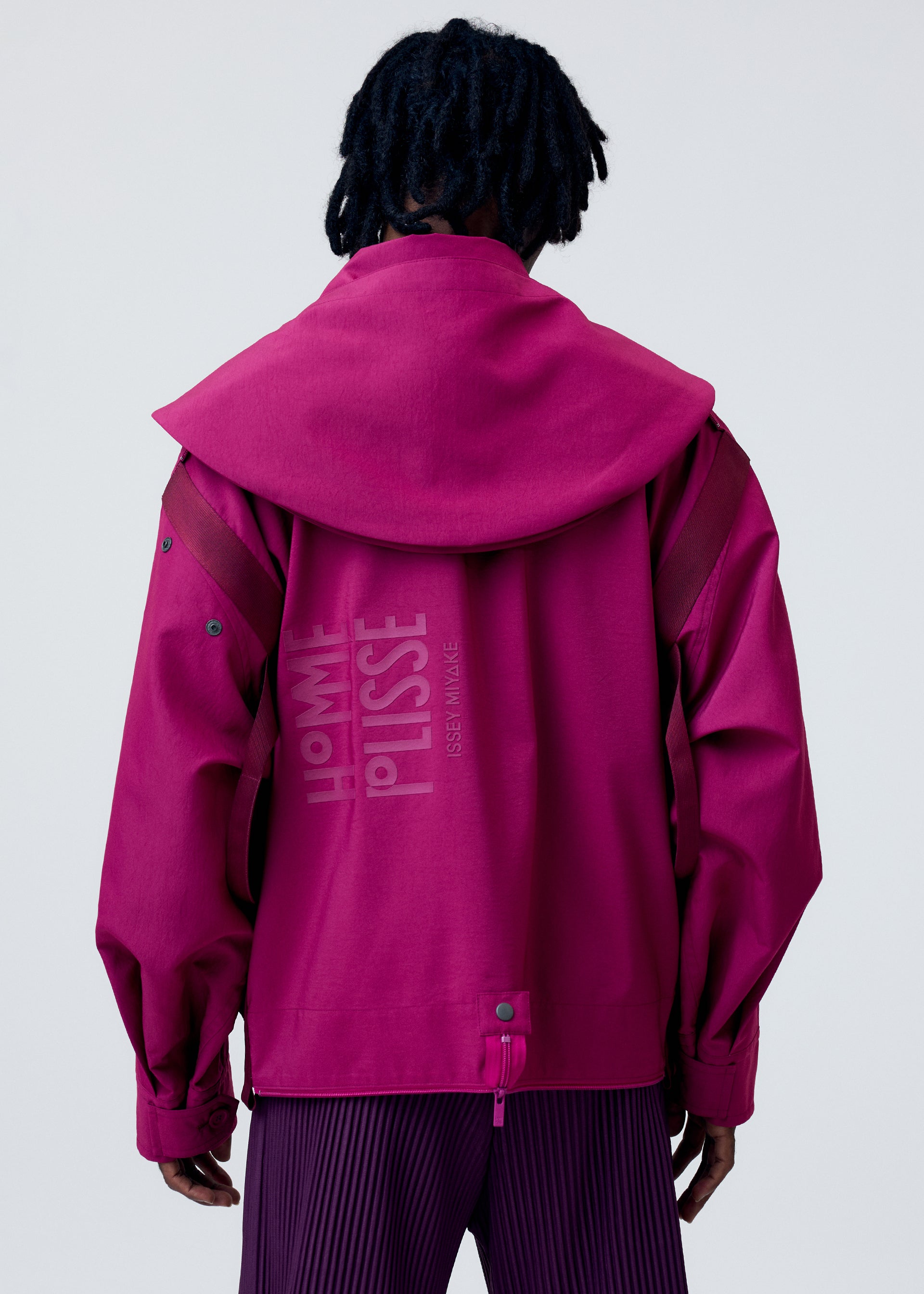 Zoomed view A person faces away, wearing the vibrant magenta CARRIER CARRIED water-repellent jacket by HOMME PLISSÉ ISSEY MIYAKE, featuring wide sleeves, an oversized collar, logo print on the back, and matching pleated pants.