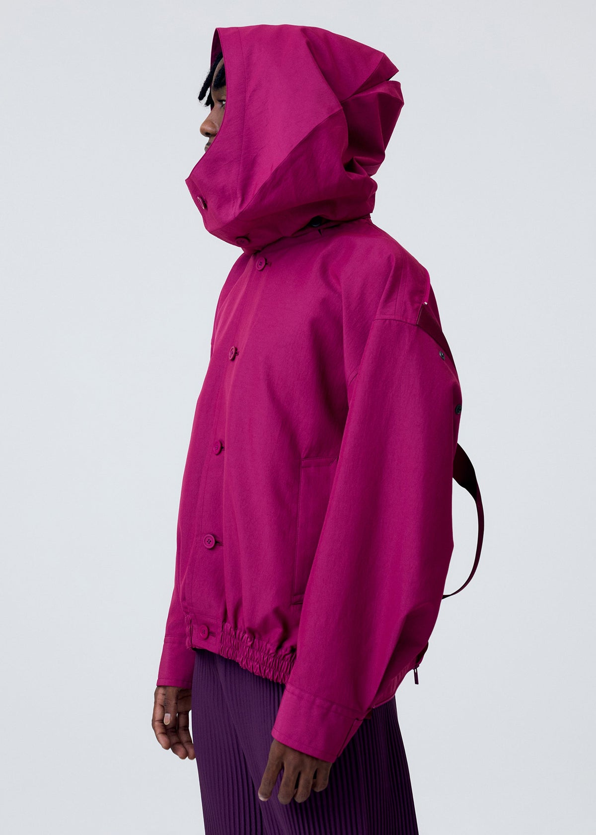 A person in profile wears HOMME PLISSÉ ISSEY MIYAKE's CARRIER CARRIED magenta water-repellent jacket with hood and a matching pleated skirt, standing against a plain light gray background. view 10 of 10