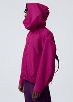A person in profile wears HOMME PLISSÉ ISSEY MIYAKE's CARRIER CARRIED magenta water-repellent jacket with hood and a matching pleated skirt, standing against a plain light gray background.