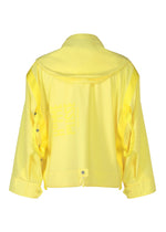 The CARRIER CARRIED jacket by HOMME PLISSÉ ISSEY MIYAKE is a bright yellow, water-repellent polyester travel piece with loose long sleeves, buttoned cuffs, side ribbon details, and “FILLE FISSE” printed vertically on the back.