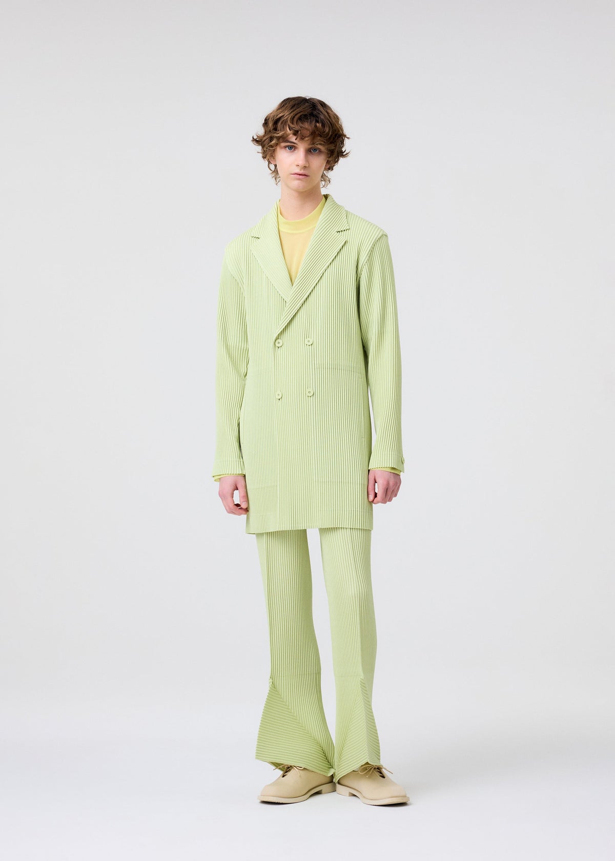 A person with curly brown hair wears the HOMME PLISSÉ ISSEY MIYAKE LINEN LIKE light green pinstripe double-breasted jacket, a yellow shirt, and cream shoes, standing against a plain white background. view 6 of 8