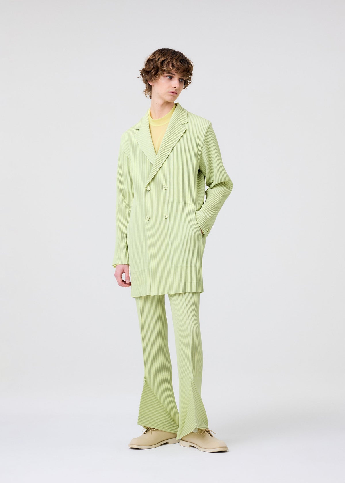 A person with curly hair wears the HOMME PLISSÉ ISSEY MIYAKE LINEN LIKE light green pinstriped suit with flared pants, double-breasted jacket, pale yellow shirt, and beige shoes, standing against a plain white background. view 7 of 8