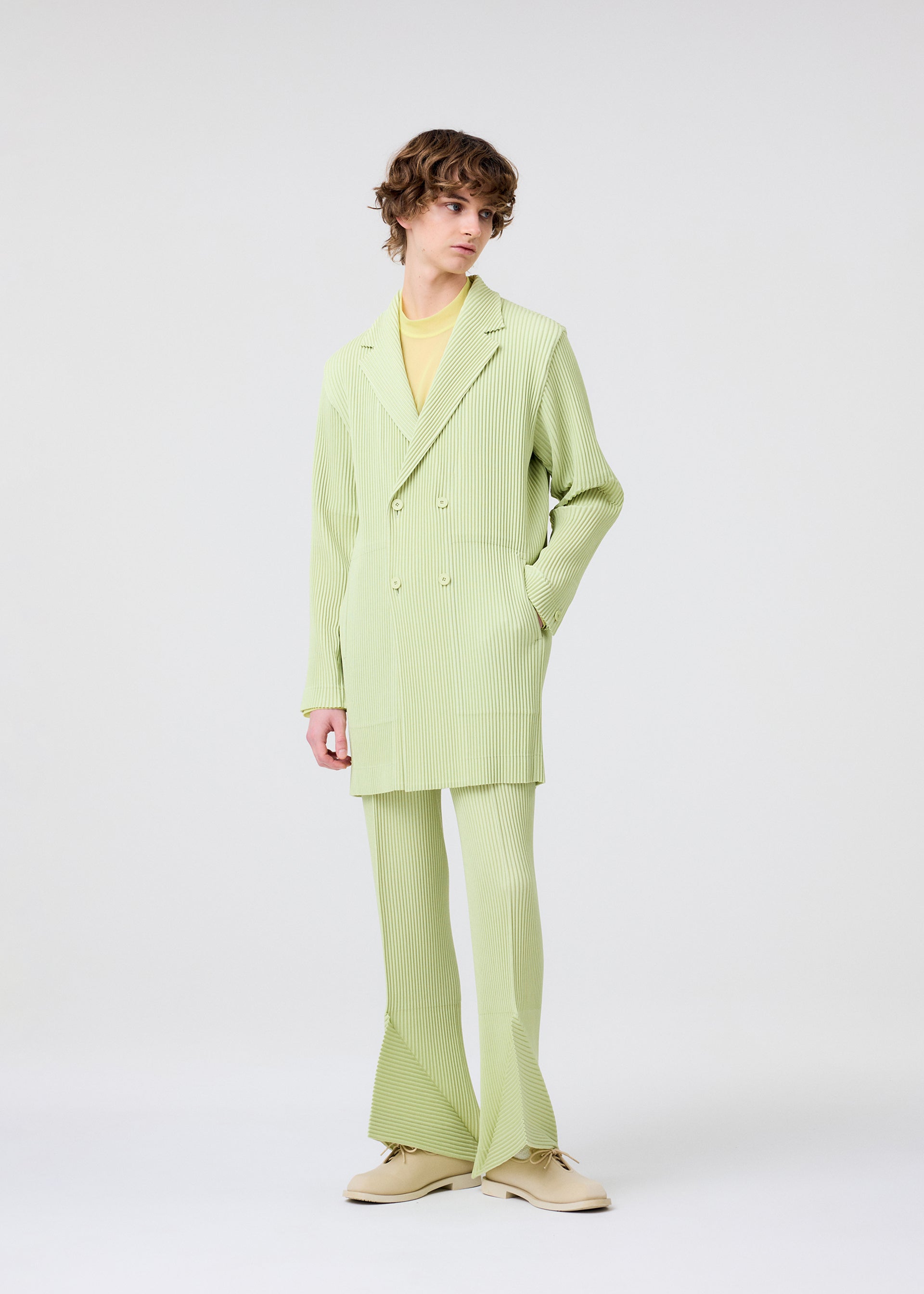 Zoomed view A person with curly hair wears the HOMME PLISSÉ ISSEY MIYAKE LINEN LIKE light green pinstriped suit with flared pants, double-breasted jacket, pale yellow shirt, and beige shoes, standing against a plain white background.