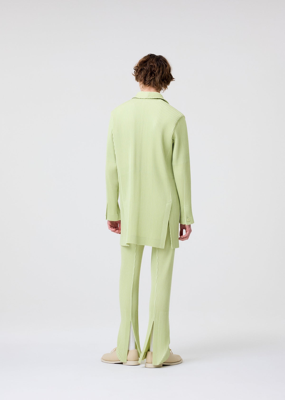 A person with short, curly hair faces away, wearing HOMME PLISSÉ ISSEY MIYAKE’s LINEN LIKE light green, long-sleeved ribbed tunic and matching ribbed pants with slit hems, creating a striking silhouette against a white background. view 8 of 8