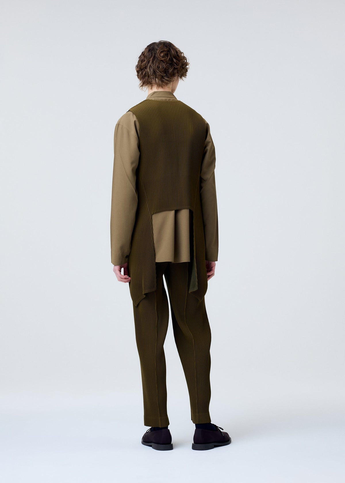 A person with curly hair faces away, wearing the olive-green TUXEDO PLEATS set from HOMME PLISSÉ ISSEY MIYAKE, featuring pleated, long-sleeved asymmetrical panels and matching pants, styled in chic layers against a light gray background. view 6 of 7