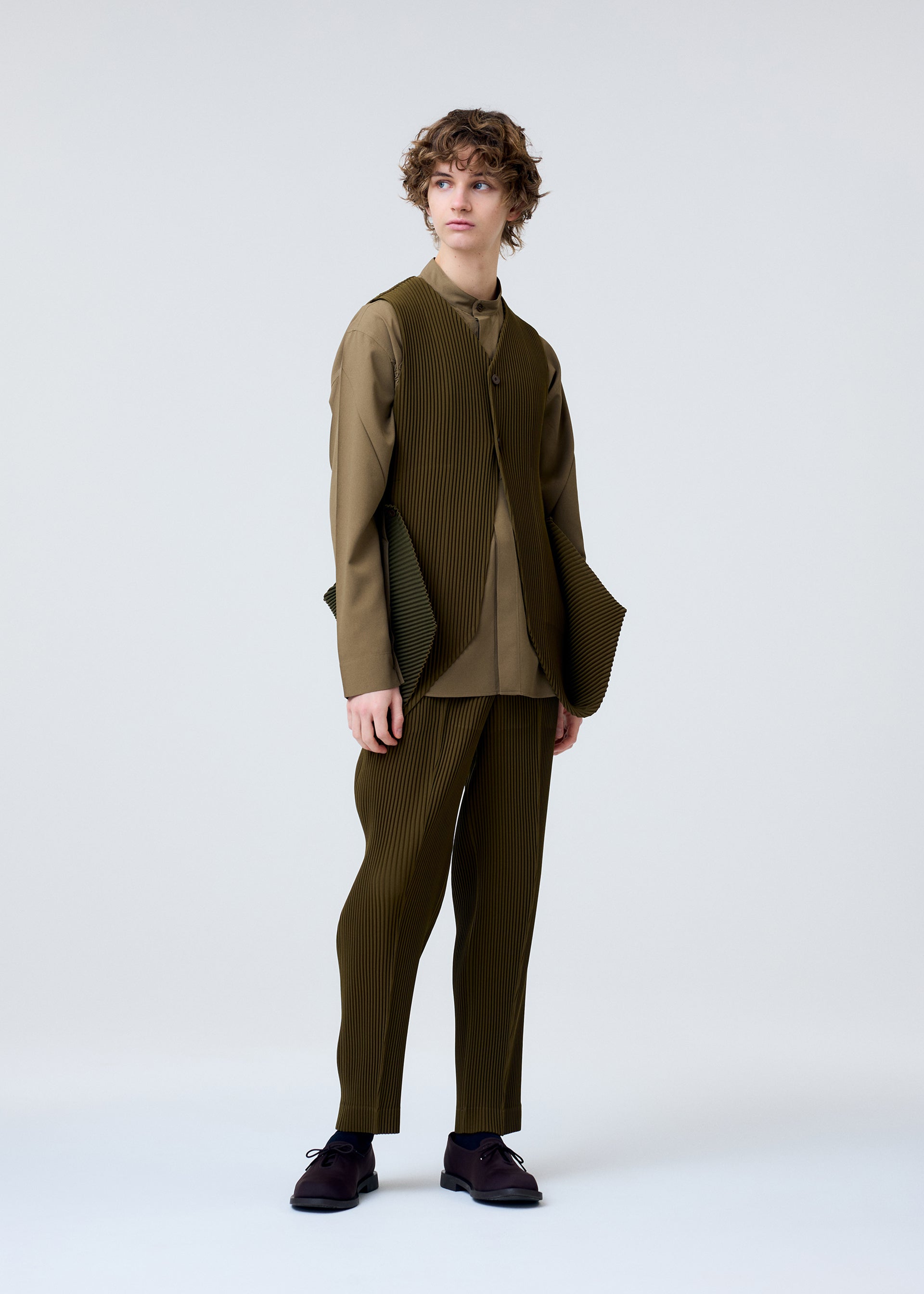 Zoomed view A person with wavy brown hair poses against a light background, wearing HOMME PLISSÉ ISSEY MIYAKE’s TUXEDO PLEATS—an olive green, loose-fit pleated suit with matching pants, a formal vest, and black dress shoes.