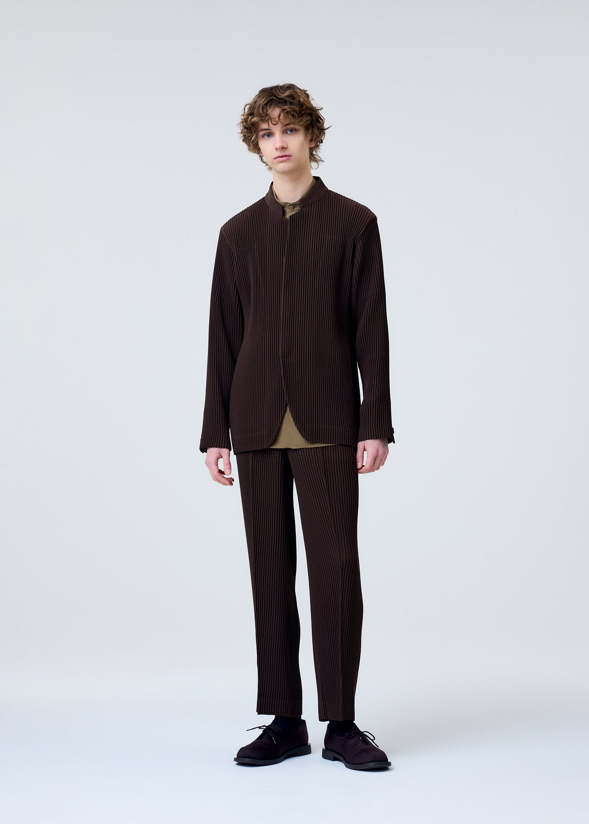 A young person with curly hair stands against a plain light background, wearing the HOMME PLISSÉ ISSEY MIYAKE TUXEDO PLEATS suit, styled with an olive-green shirt and black shoes. They face forward with a neutral expression and relaxed posture. view 7 of 9