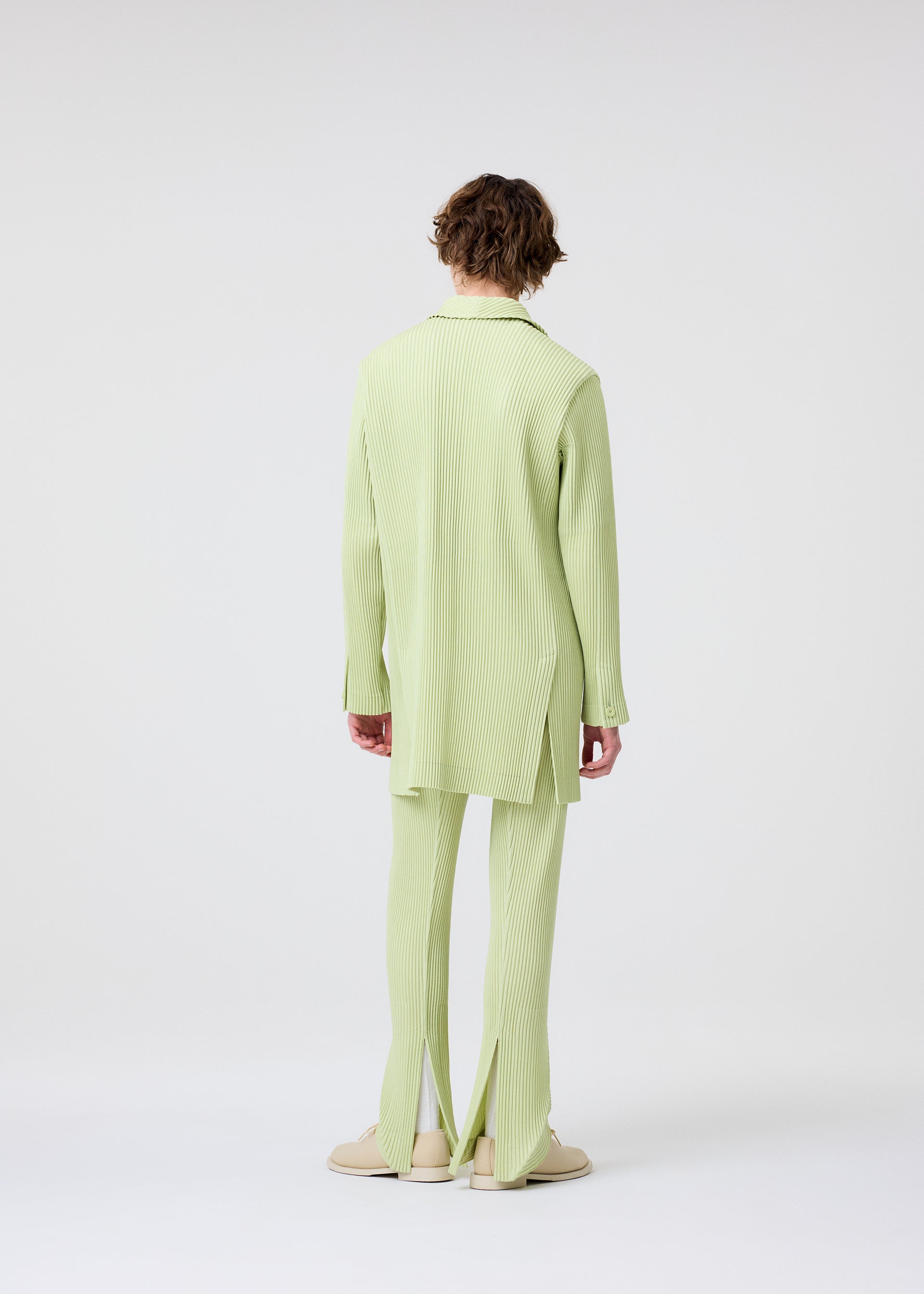 Zoomed view A person with short, curly brown hair faces away, wearing HOMME PLISSÉ ISSEY MIYAKE's LINEN LIKE light green ribbed long blazer and linen-texture pants with slit hems, paired with beige shoes against a plain white background.