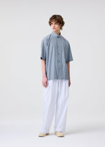 A person with curly hair models the FRESHEN shirt by HOMME PLISSÉ ISSEY MIYAKE— a loose, short-sleeve blue and white striped top with a wrinkled texture—paired with wide white pants and cream shoes against a plain white background.