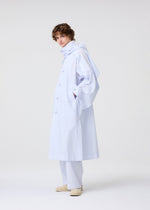 A person with short curly hair stands sideways in HOMME PLISSÉ ISSEY MIYAKE’s FRESHEN: a loose, light blue trench coat with subtle wrinkles over matching wide pants and cream shoes, against a plain white background.