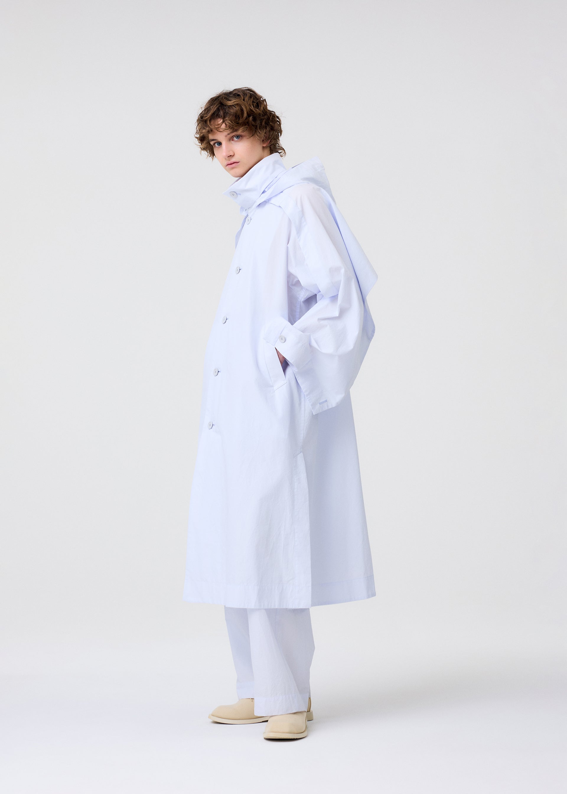 Zoomed view A person with short curly hair stands sideways in HOMME PLISSÉ ISSEY MIYAKE’s FRESHEN: a loose, light blue trench coat with subtle wrinkles over matching wide pants and cream shoes, against a plain white background.