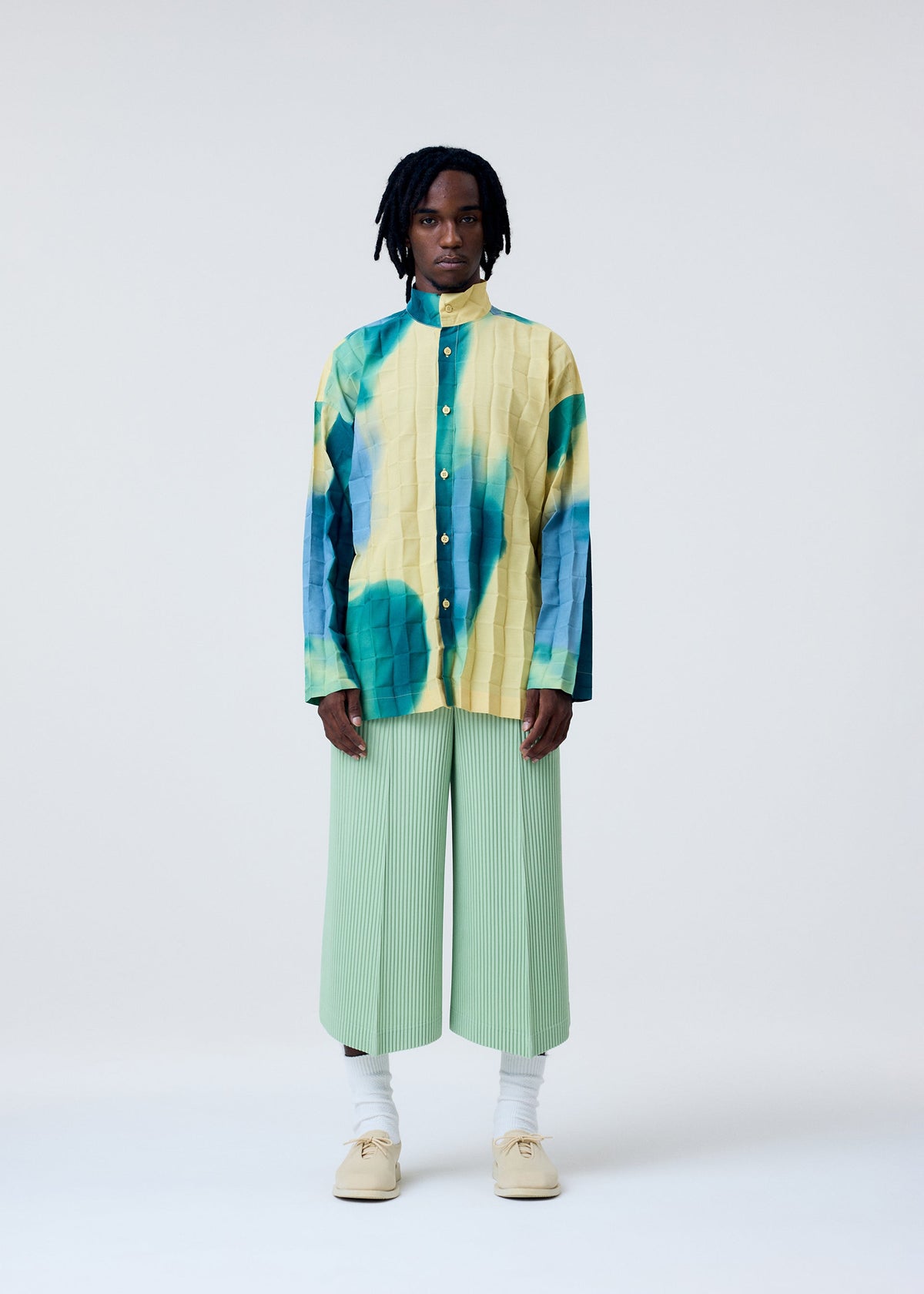 A person stands against a plain background wearing the HOMME PLISSÉ ISSEY MIYAKE DAWN EDGE SHIRT, light green hand-pleated wide-leg pants, white socks, and beige shoes. view 7 of 10