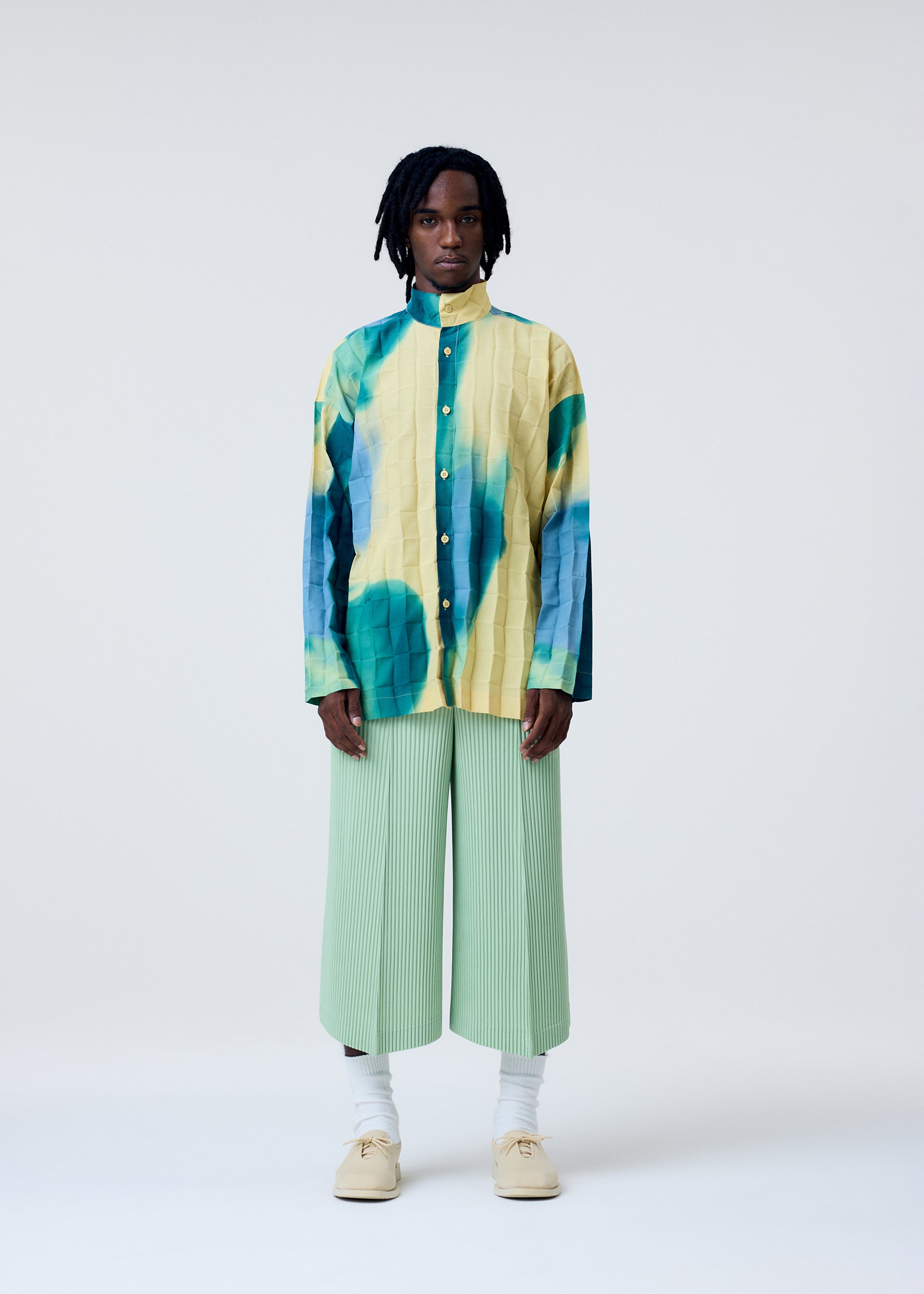 Zoomed view A person stands against a plain background wearing the HOMME PLISSÉ ISSEY MIYAKE DAWN EDGE SHIRT, light green hand-pleated wide-leg pants, white socks, and beige shoes.