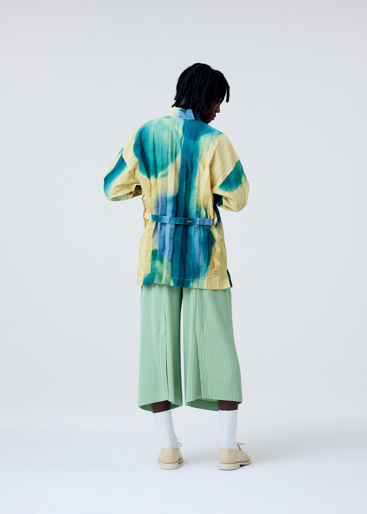 Against a plain light background, a person faces away wearing the HOMME PLISSÉ ISSEY MIYAKE DAWN EDGE SHIRT, a loose yellow jacket with blue-green streaks and Nassen print over a standing collar shirt, paired with light green wide-legged pants. view 8 of 10