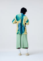 Against a plain light background, a person faces away wearing the HOMME PLISSÉ ISSEY MIYAKE DAWN EDGE SHIRT, a loose yellow jacket with blue-green streaks and Nassen print over a standing collar shirt, paired with light green wide-legged pants.