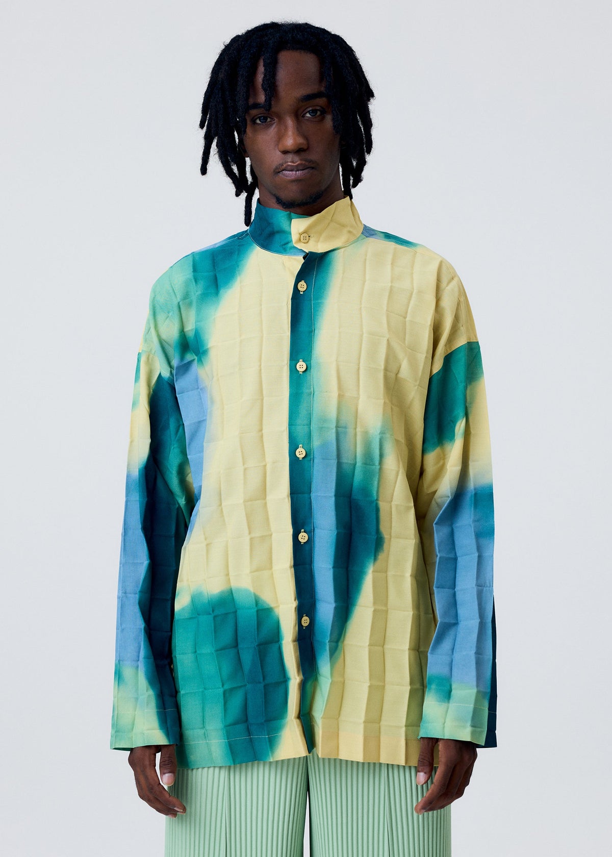 A person with medium-length dreadlocks wears the DAWN EDGE SHIRT by HOMME PLISSÉ ISSEY MIYAKE—a loose, long-sleeve standing collar shirt with a yellow and teal tie-dye pattern—paired with light green hand-pleated pants against a plain light background. view 9 of 10