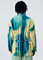 Seen from behind, a person with medium-length dreadlocks wears the HOMME PLISSÉ ISSEY MIYAKE DAWN EDGE SHIRT—a colorful, grid-patterned top in yellow, teal, and green—paired with a matching belt and pleated green pants.