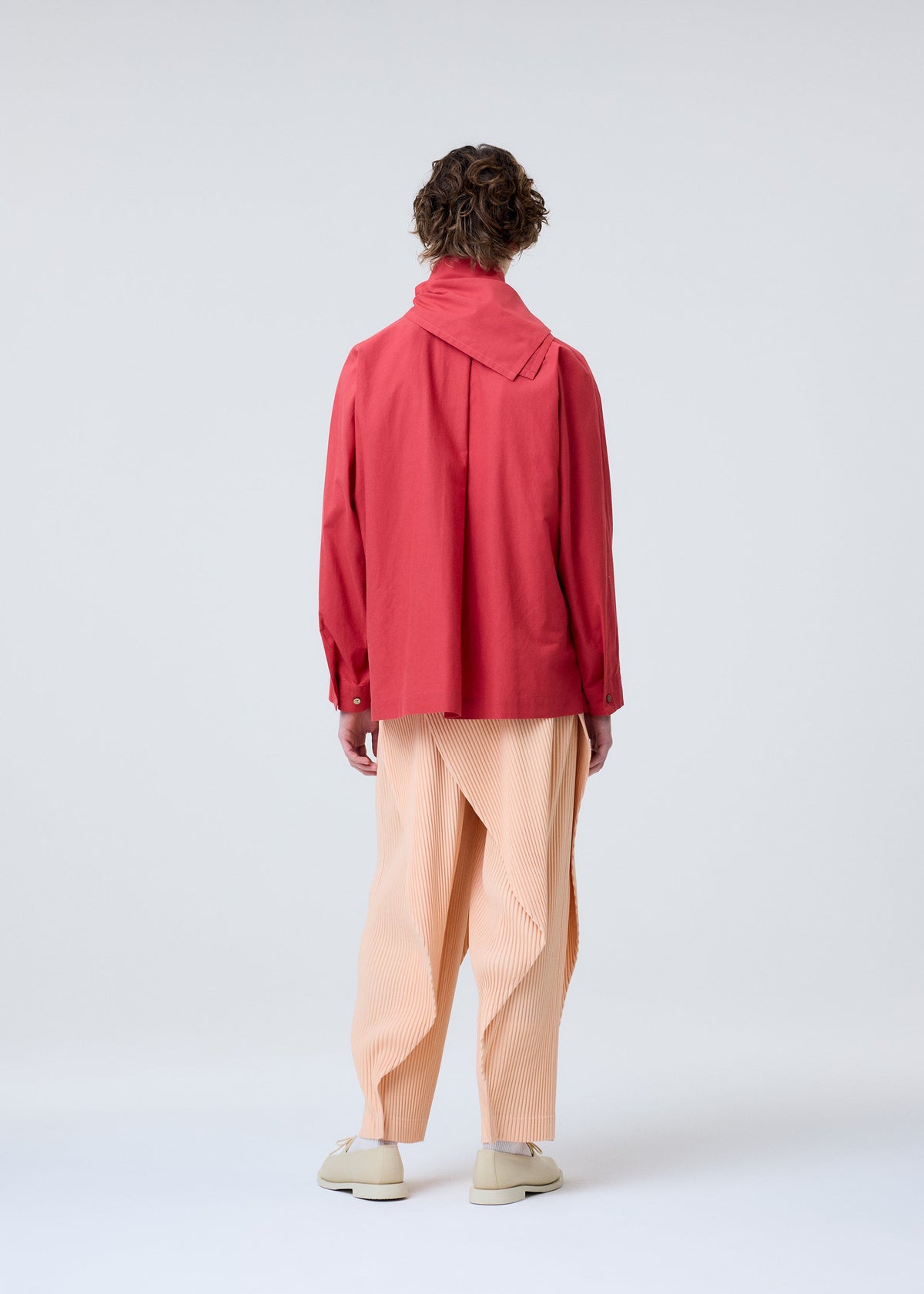 A person with short curly hair wears the HOMME PLISSÉ ISSEY MIYAKE STREAMLINE SHIRT, featuring a loose organic cotton fit and wide scarf neckline, plus peach pleated trousers and light shoes. They stand against a plain light gray background. view 6 of 7