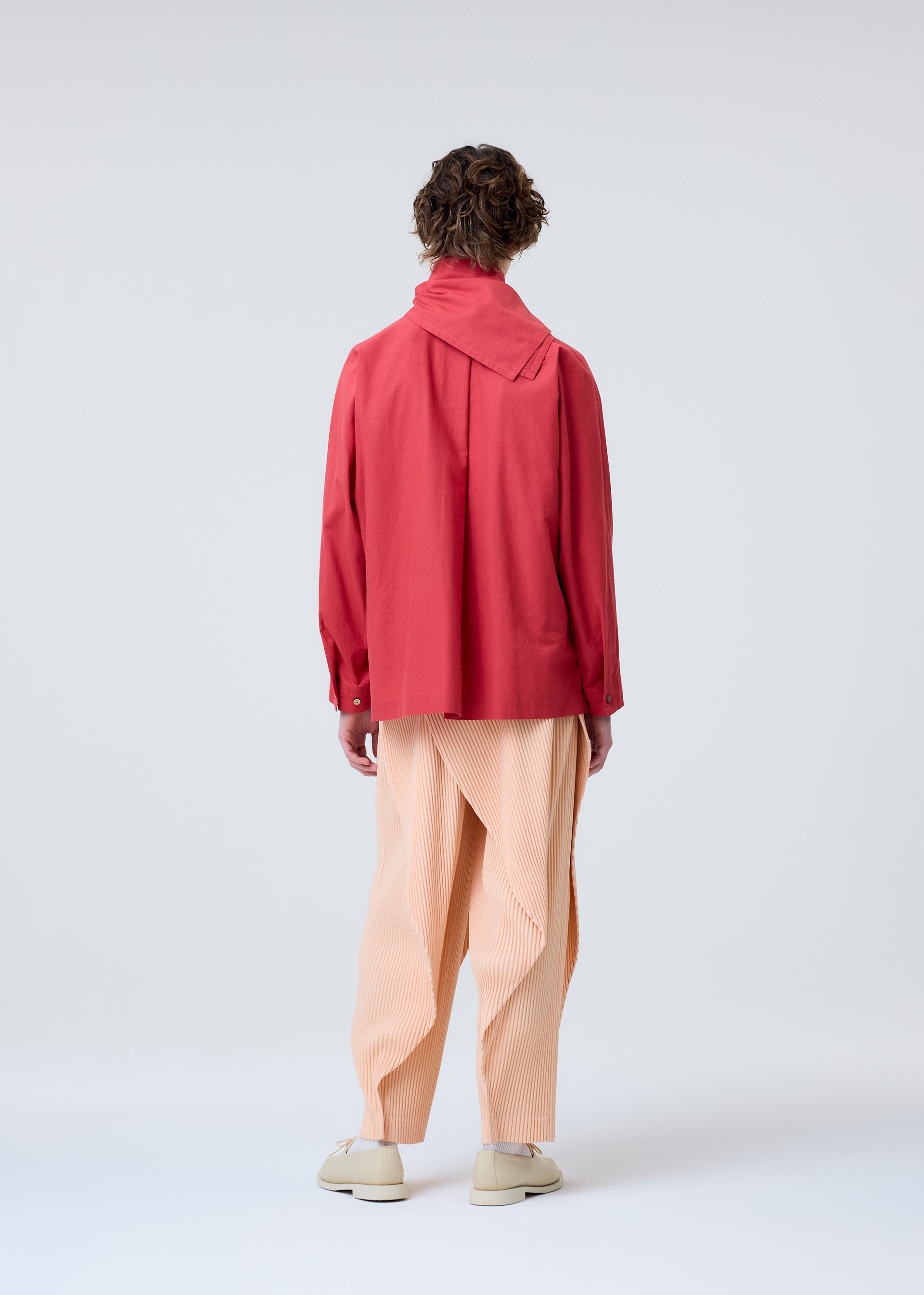 Zoomed view A person with short curly hair wears the HOMME PLISSÉ ISSEY MIYAKE STREAMLINE SHIRT, featuring a loose organic cotton fit and wide scarf neckline, plus peach pleated trousers and light shoes. They stand against a plain light gray background.