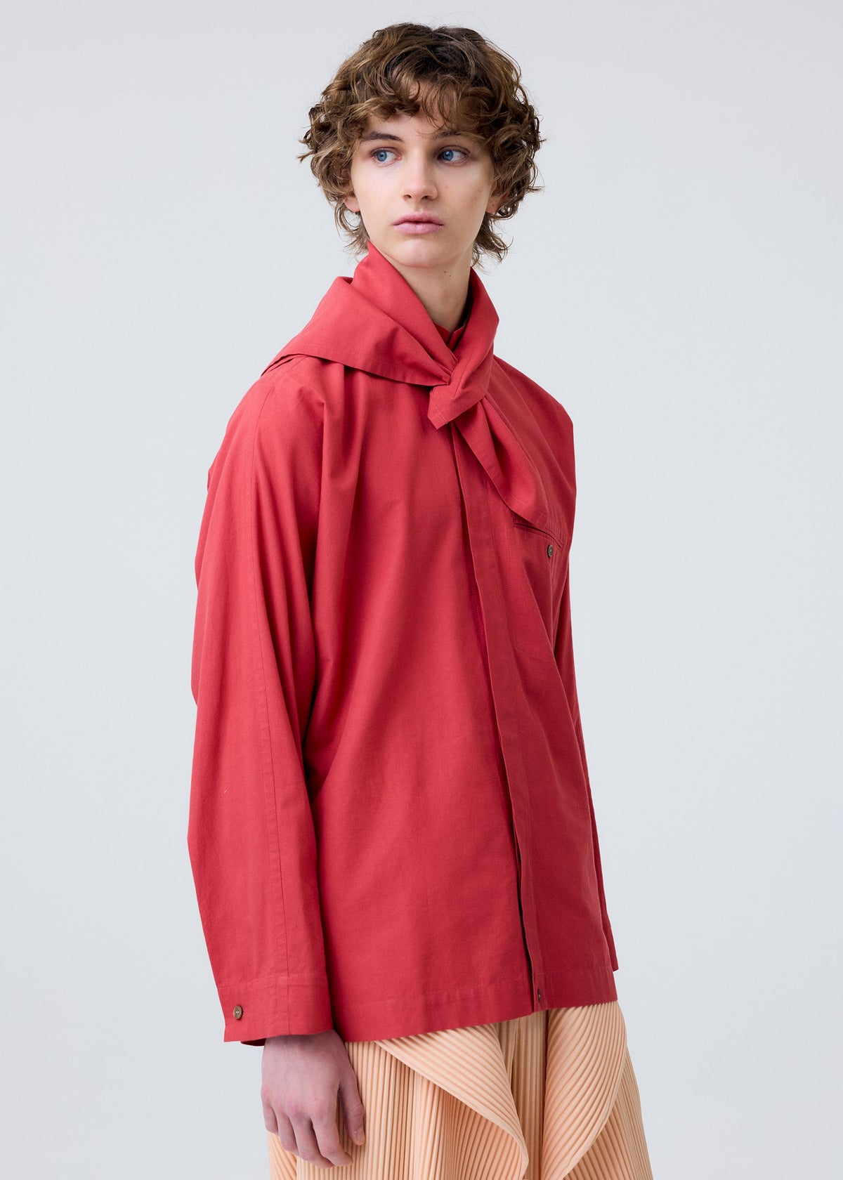 A person with short curly hair wears the HOMME PLISSÉ ISSEY MIYAKE STREAMLINE SHIRT in red, featuring a scarf neckline, and a pleated beige skirt, standing against a plain light background and gazing slightly to the side. view 7 of 7
