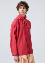 A person with short curly hair wears the HOMME PLISSÉ ISSEY MIYAKE STREAMLINE SHIRT in red, featuring a scarf neckline, and a pleated beige skirt, standing against a plain light background and gazing slightly to the side.