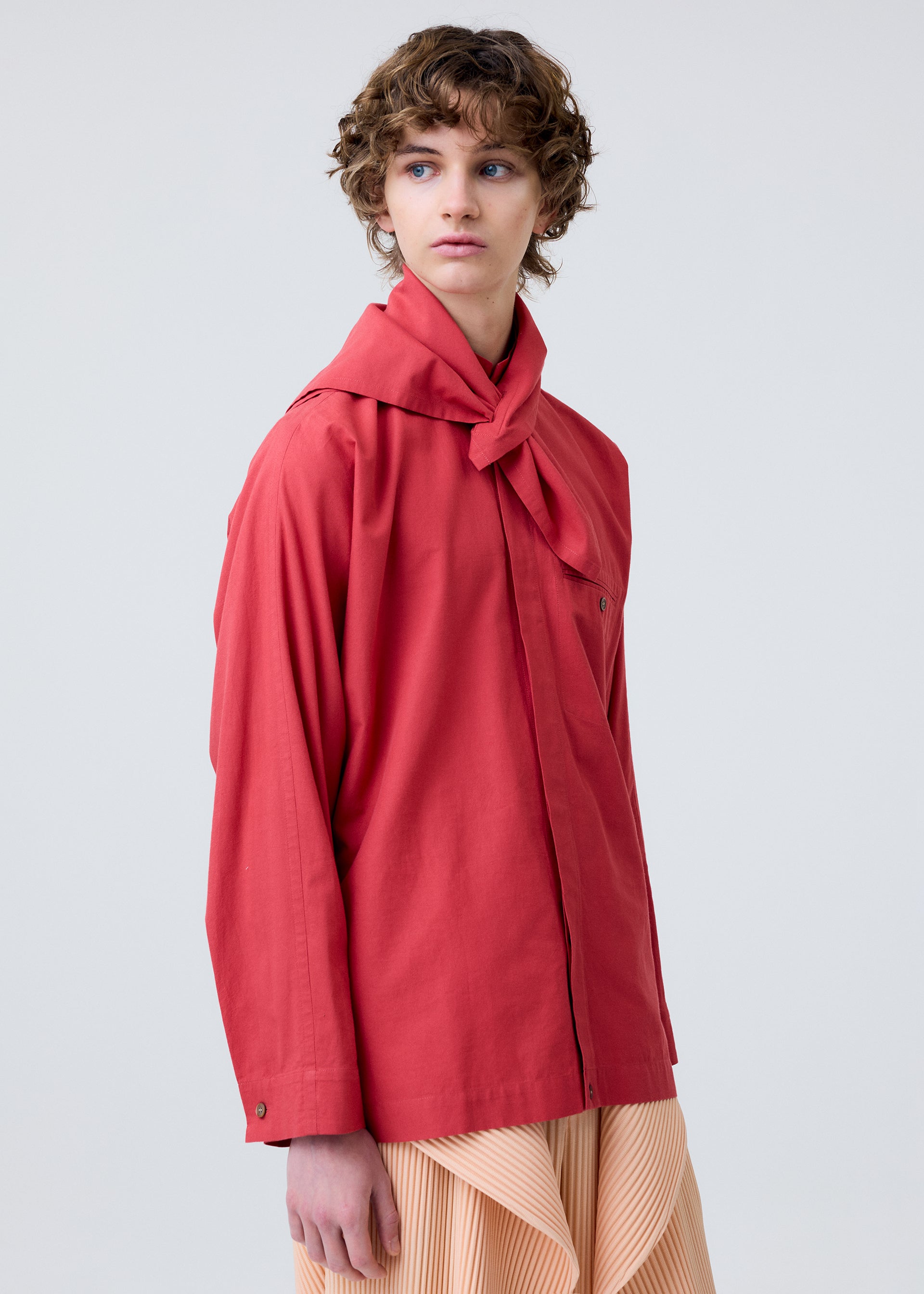 Zoomed view A person with short curly hair wears the HOMME PLISSÉ ISSEY MIYAKE STREAMLINE SHIRT in red, featuring a scarf neckline, and a pleated beige skirt, standing against a plain light background and gazing slightly to the side.