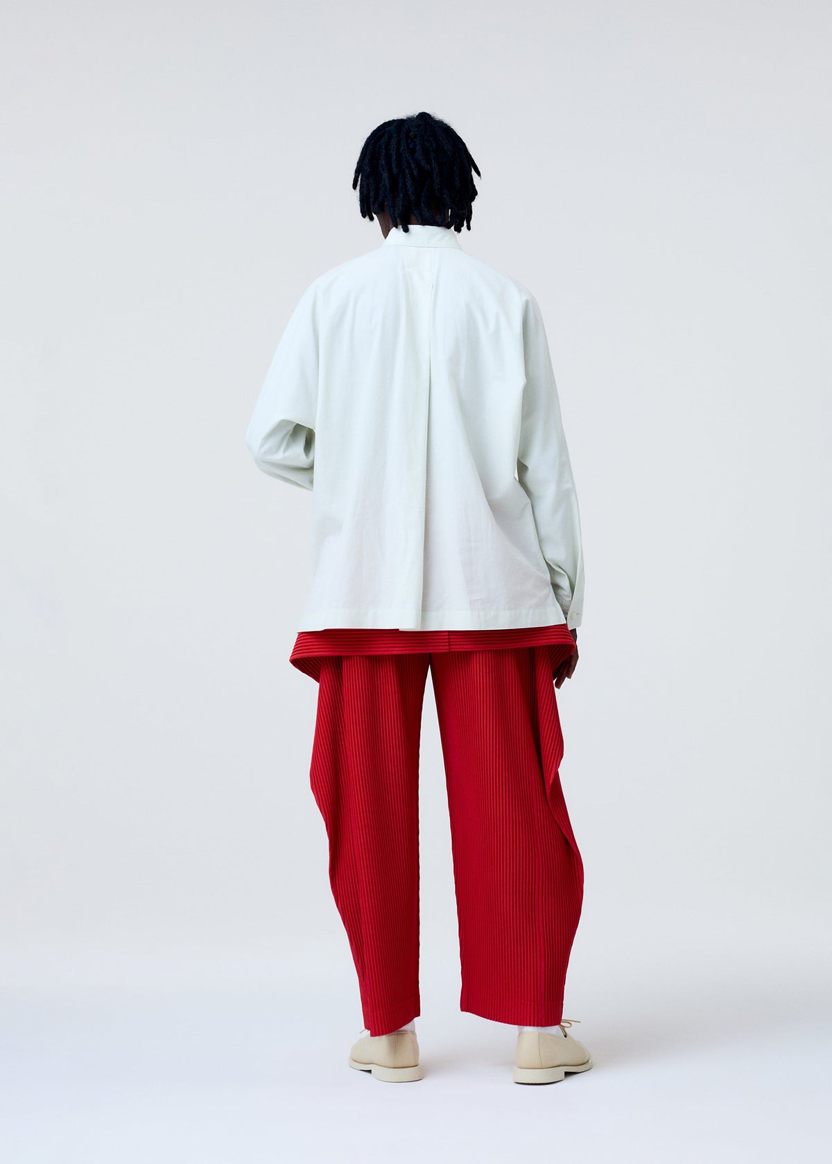 A person faces away from the camera, wearing the HOMME PLISSÉ ISSEY MIYAKE STREAMLINE SHIRT and red pleated pants with draped panels, paired with cream shoes. The background is plain white. view 7 of 8