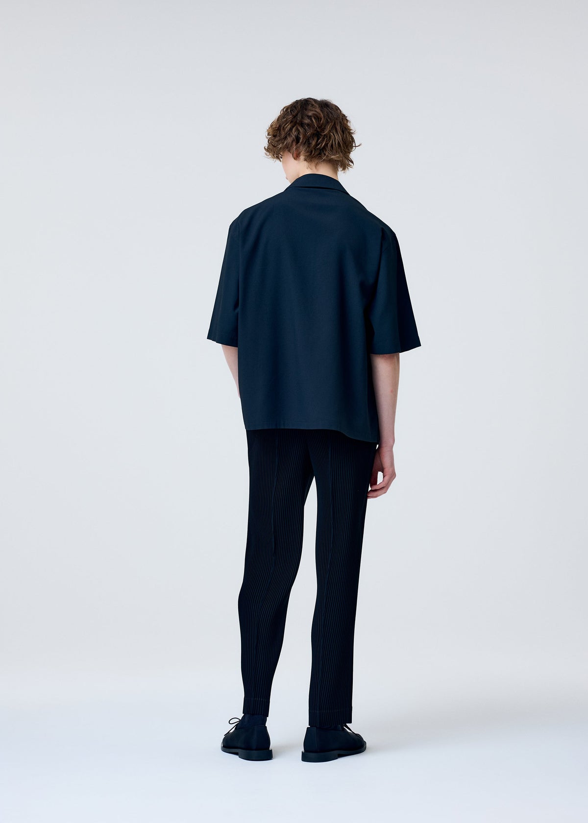 A person with short, wavy hair stands against a plain light background, facing away. They are wearing the EMBROIDERY PRESS SHIRT by HOMME PLISSÉ ISSEY MIYAKE, paired with dark pinstripe pants and black shoes. view 7 of 9