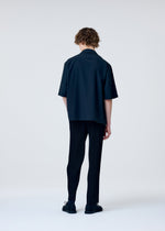A person with short, wavy hair stands against a plain light background, facing away. They are wearing the EMBROIDERY PRESS SHIRT by HOMME PLISSÉ ISSEY MIYAKE, paired with dark pinstripe pants and black shoes.