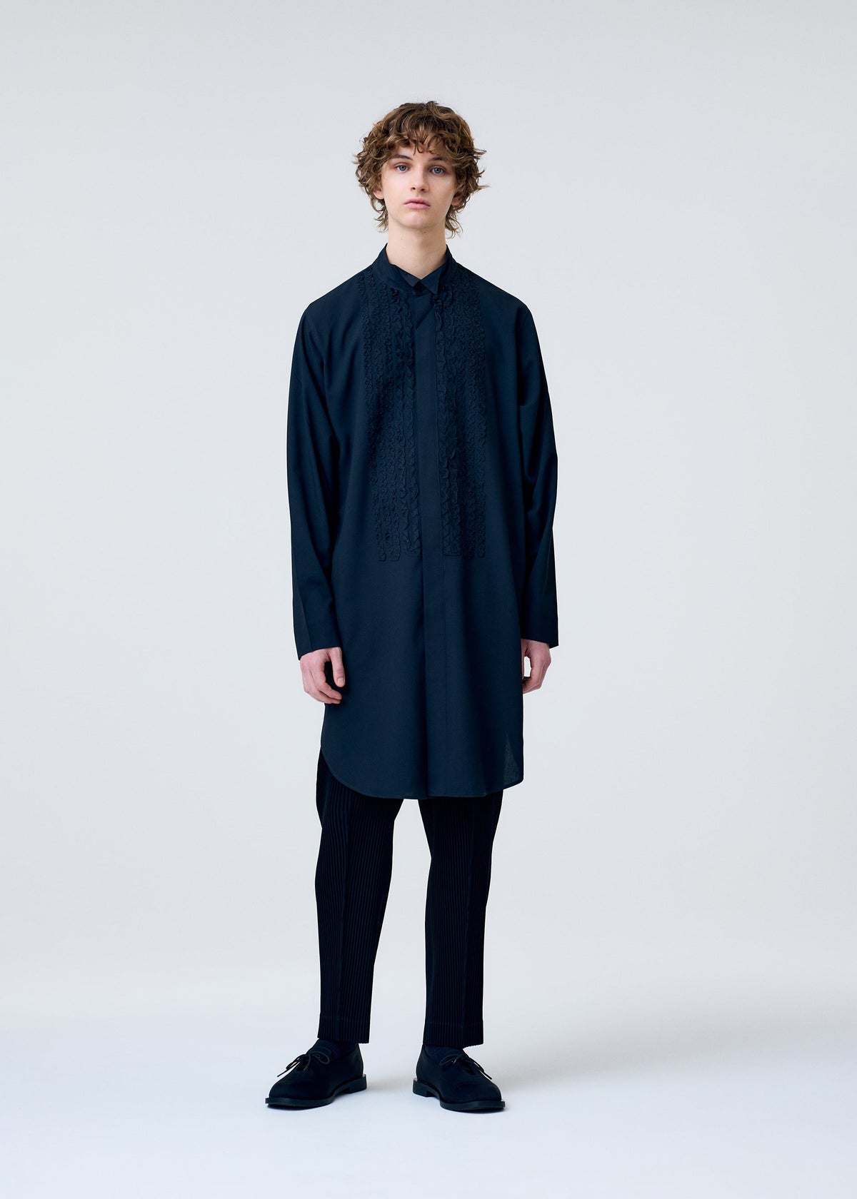 A person with curly hair stands against a light background, wearing the EMBROIDERY PRESS SHIRT by HOMME PLISSÉ ISSEY MIYAKE under a dark tunic, styled with black pinstriped trousers and shoes for a sleek, minimalist look. view 6 of 8