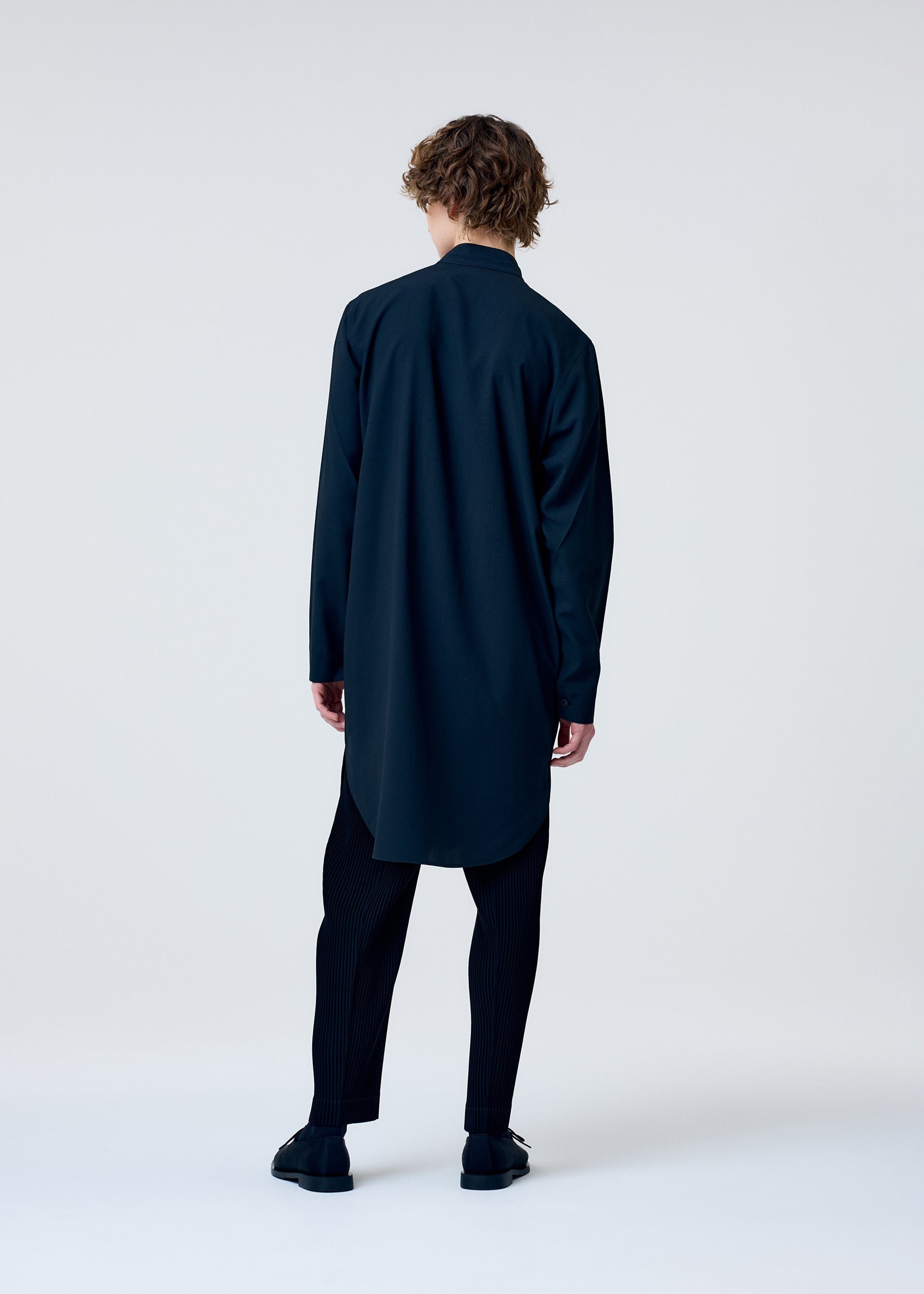 Zoomed view A person with curly hair faces away from the camera, wearing the EMBROIDERY PRESS SHIRT by HOMME PLISSÉ ISSEY MIYAKE, paired with black pants and shoes against a plain light background.