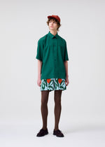 A person wears the green PL RAMIE SHIRT by HOMME PLISSÉ ISSEY MIYAKE, patterned shorts with orange and green shapes, brown tights and shoes, and a red cap against a plain light background.