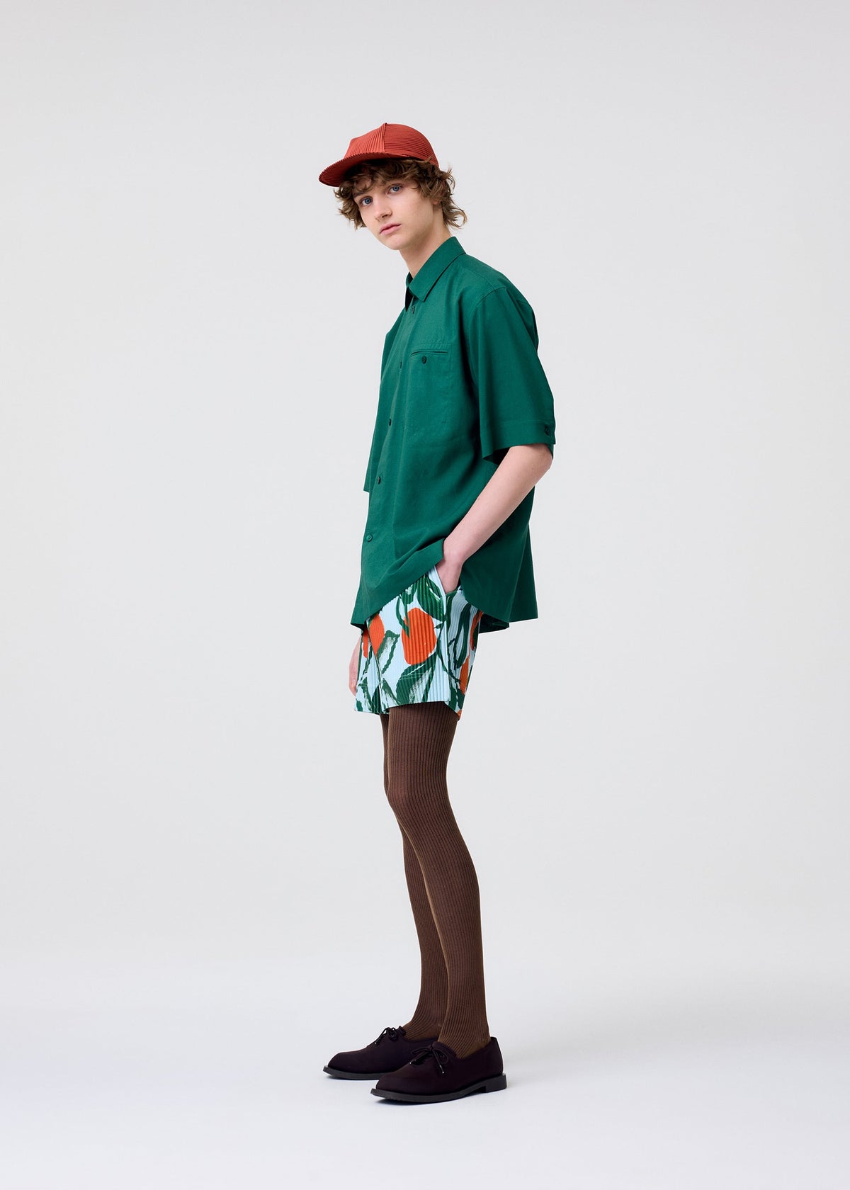 A person with curly hair and a neutral expression stands against a plain background, wearing the HOMME PLISSÉ ISSEY MIYAKE PL RAMIE SHIRT in green, colorful patterned shorts, brown tights, black shoes, and a red cap. view 7 of 8