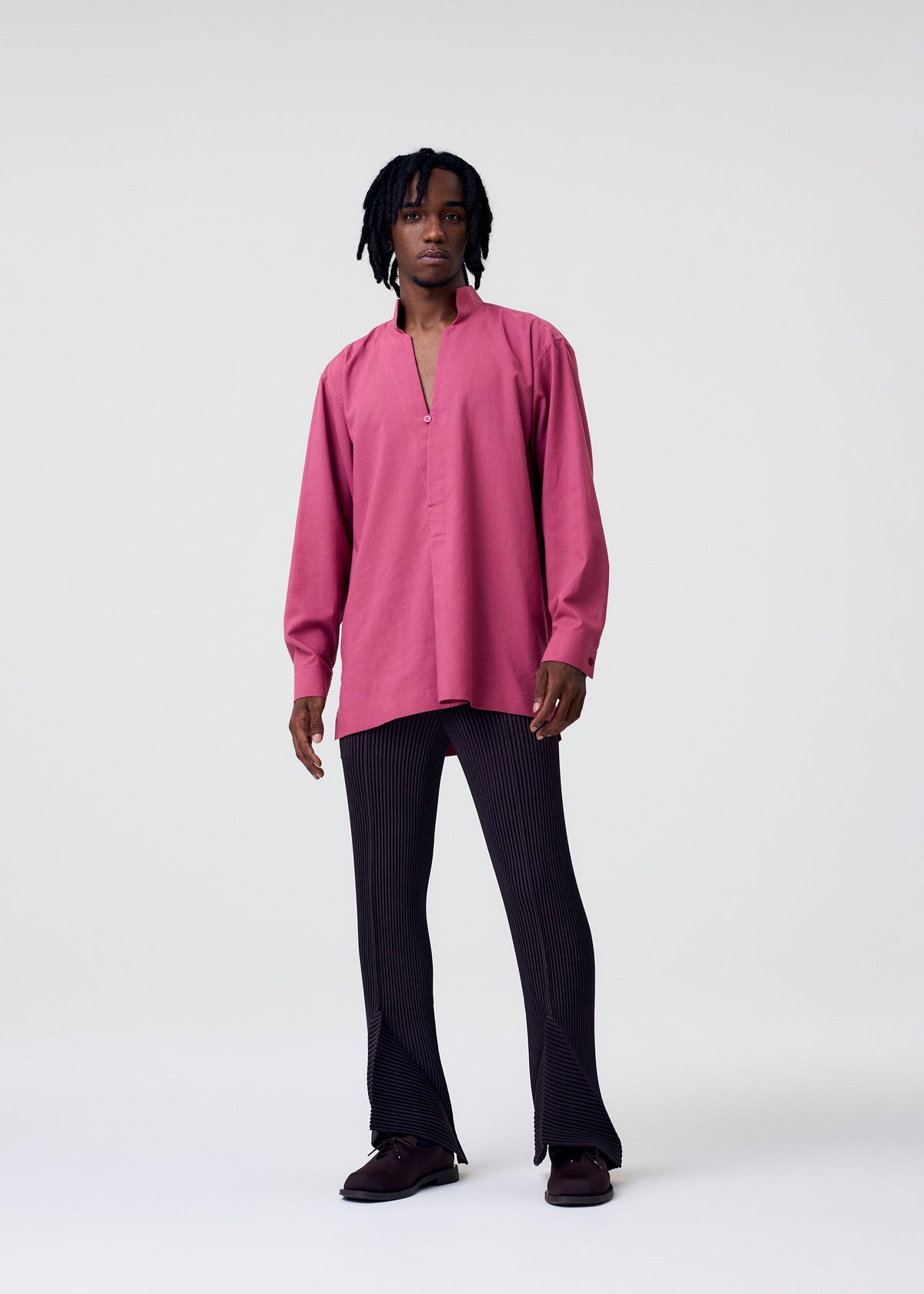 Someone with short dreadlocks wears the HOMME PLISSÉ ISSEY MIYAKE PL RAMIE SHIRT—a loose, wrinkle-resistant magenta collared top with a partial front opening—paired with black flared pants and dark shoes against a plain light background. view 6 of 7