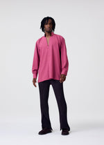 Someone with short dreadlocks wears the HOMME PLISSÉ ISSEY MIYAKE PL RAMIE SHIRT—a loose, wrinkle-resistant magenta collared top with a partial front opening—paired with black flared pants and dark shoes against a plain light background.