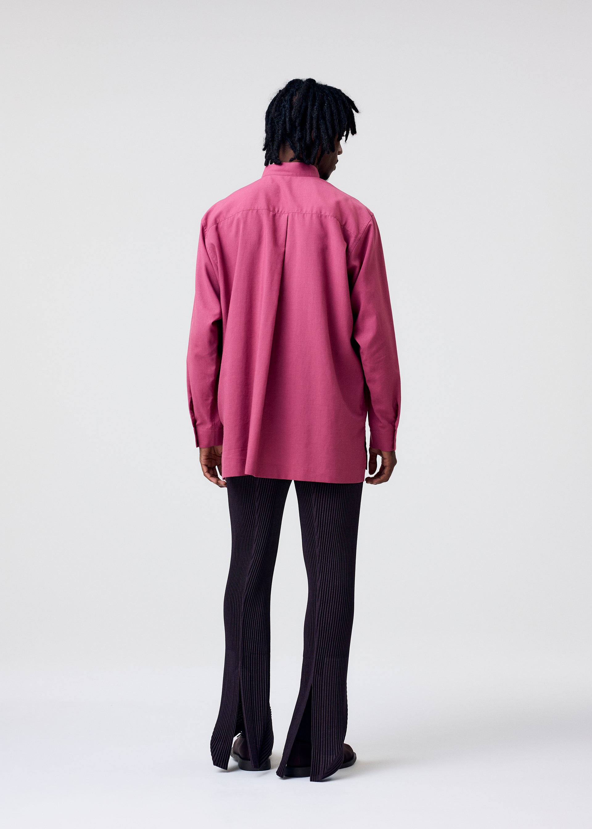 Zoomed view A person with dark braided hair stands facing away, wearing the HOMME PLISSÉ ISSEY MIYAKE PL RAMIE SHIRT in magenta and dark, pinstriped pants with back slits, set against a plain light background.