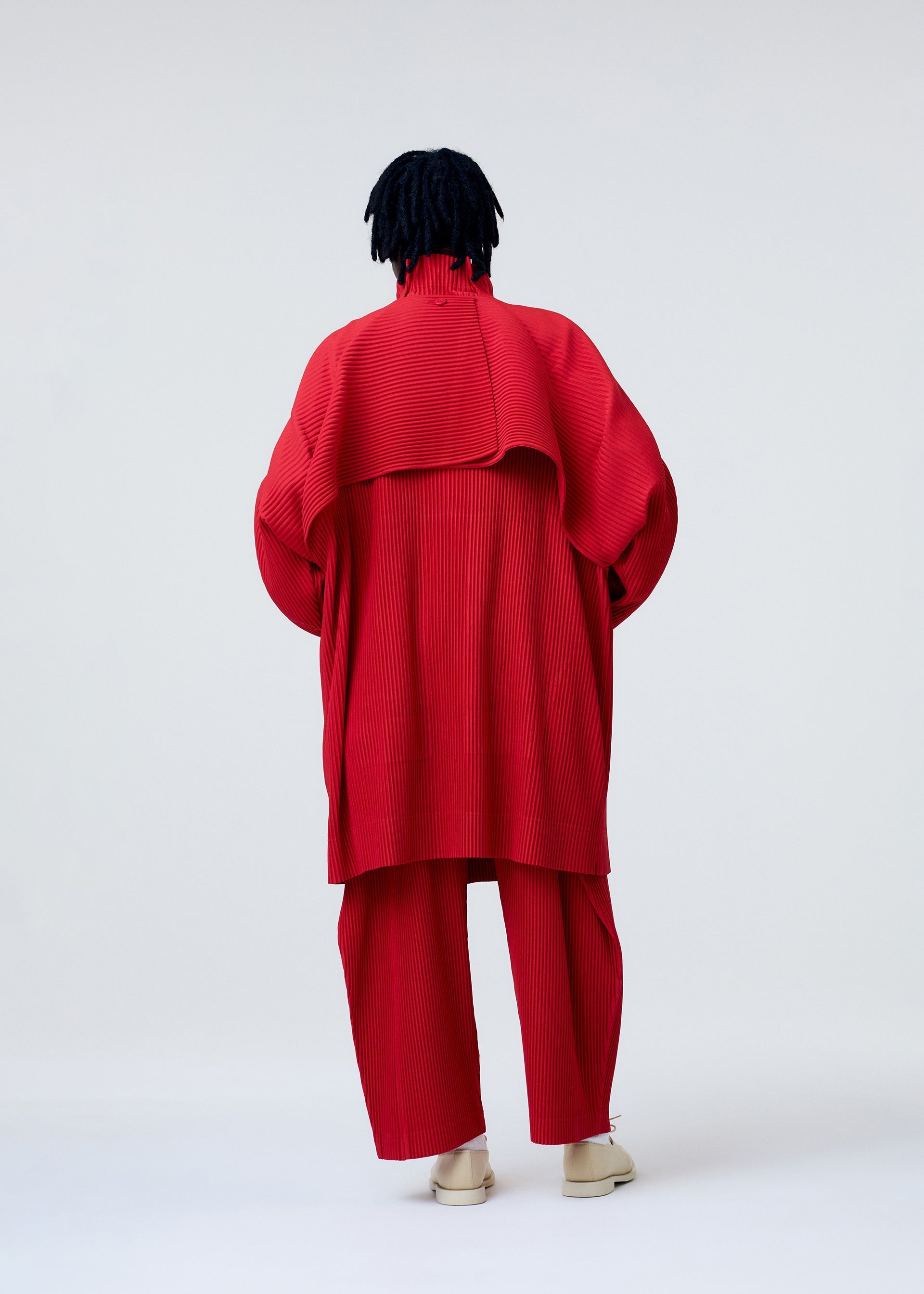 Zoomed view A person with short dreadlocks stands back to the camera, wearing HOMME PLISSÉ ISSEY MIYAKE’s COVER AND FOLD architectural red coat with 3D pleating and white shoes, set against a plain light background.