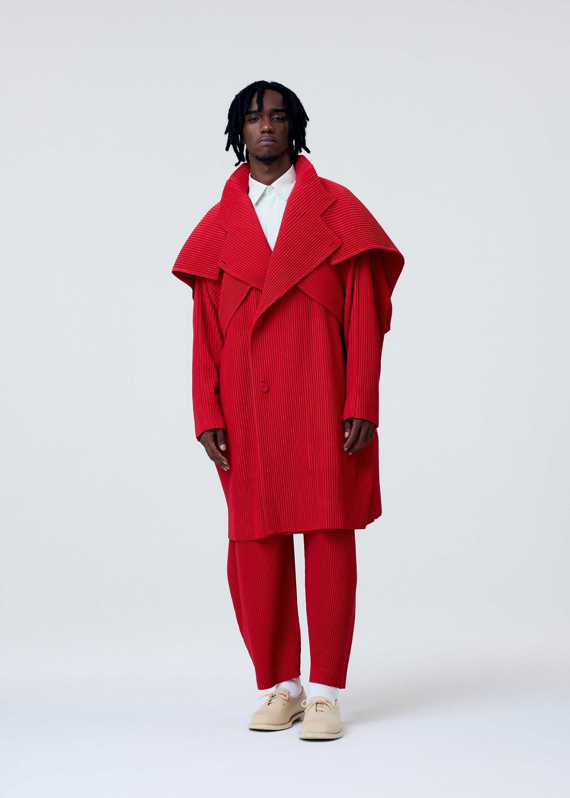 Zoomed view A person stands against a plain background wearing the COVER AND FOLD coat and pants from HOMME PLISSÉ ISSEY MIYAKE, featuring bright red three-dimensional pleating, styled with a white collared shirt and cream shoes.
