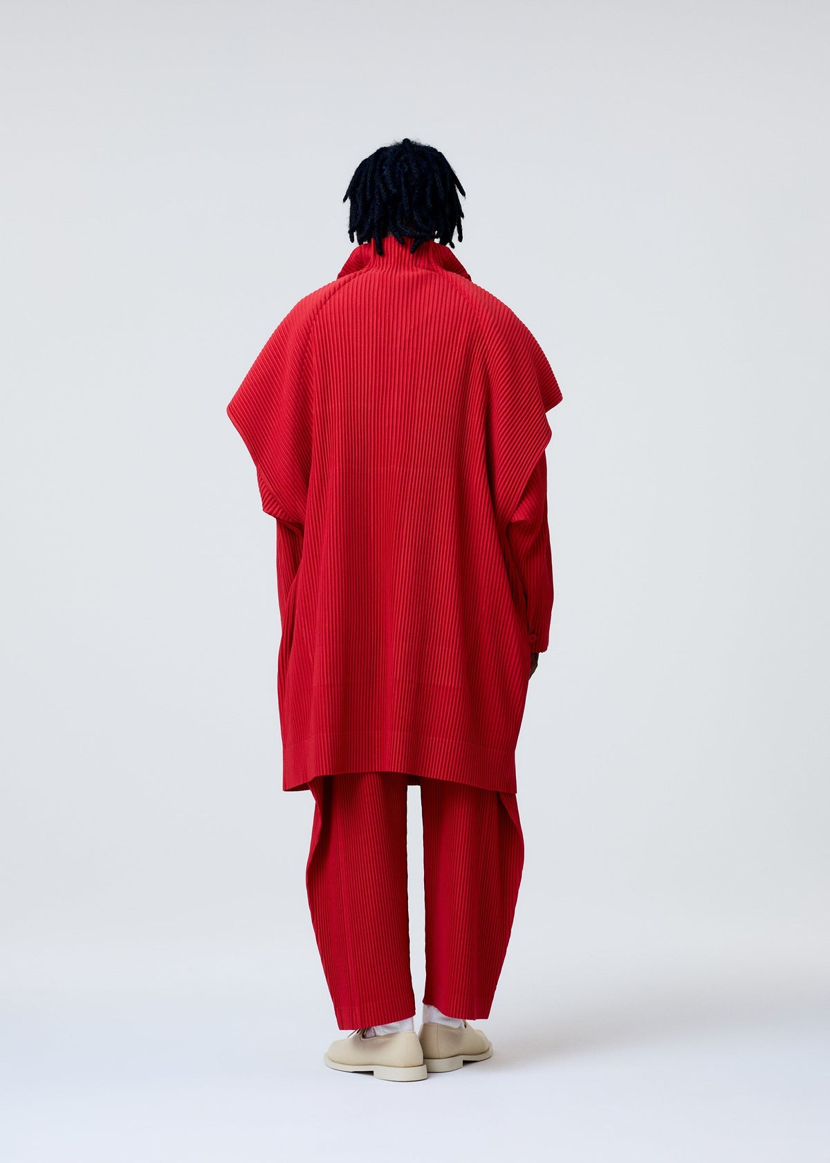 A person with dark, textured hair faces away, wearing the bright red COVER AND FOLD outfit by HOMME PLISSÉ ISSEY MIYAKE, with three-dimensional pleating, wide sleeves, matching pants, and light shoes against a plain light background. view 6 of 6
