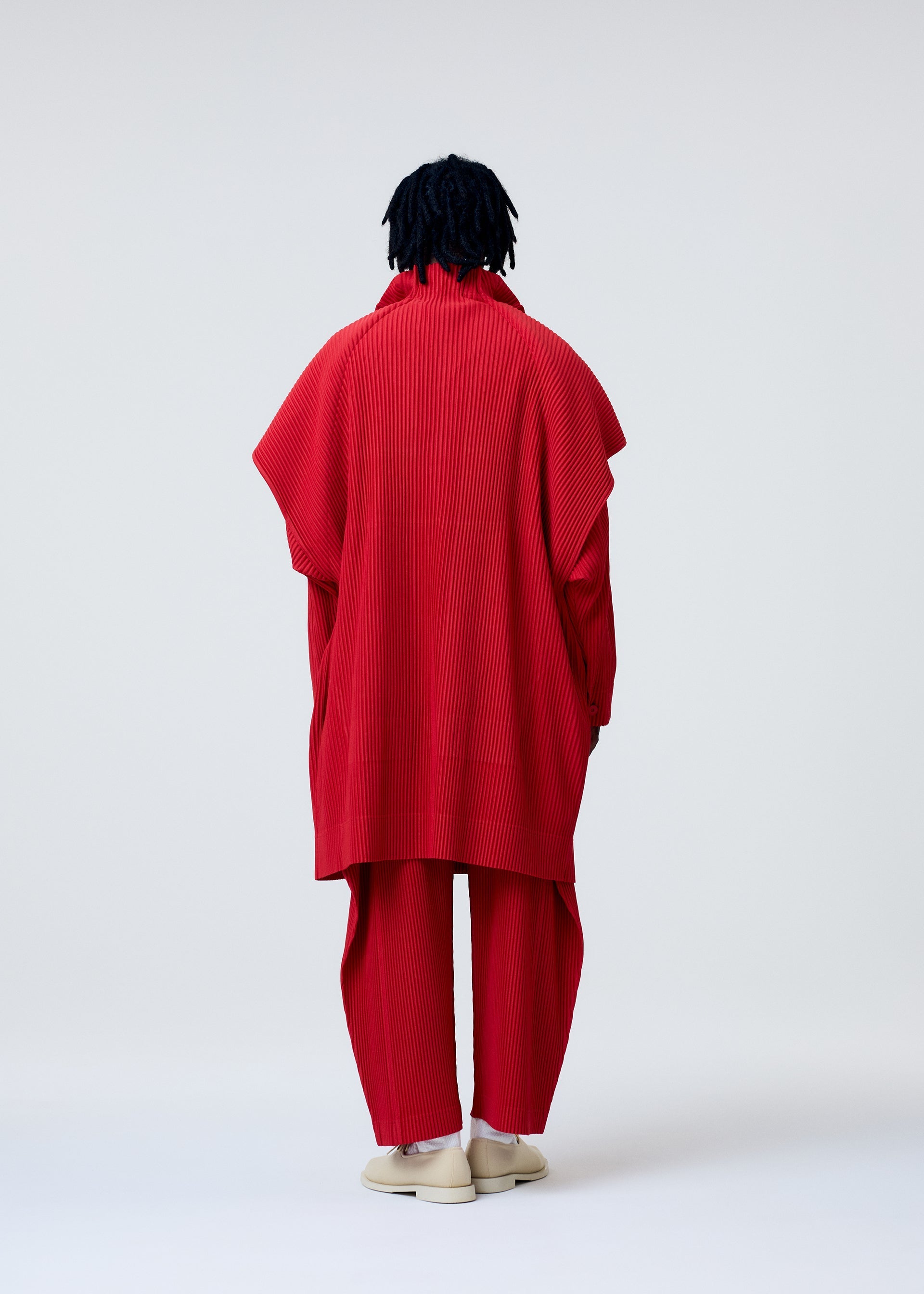 Zoomed view A person with dark, textured hair faces away, wearing the bright red COVER AND FOLD outfit by HOMME PLISSÉ ISSEY MIYAKE, with three-dimensional pleating, wide sleeves, matching pants, and light shoes against a plain light background.