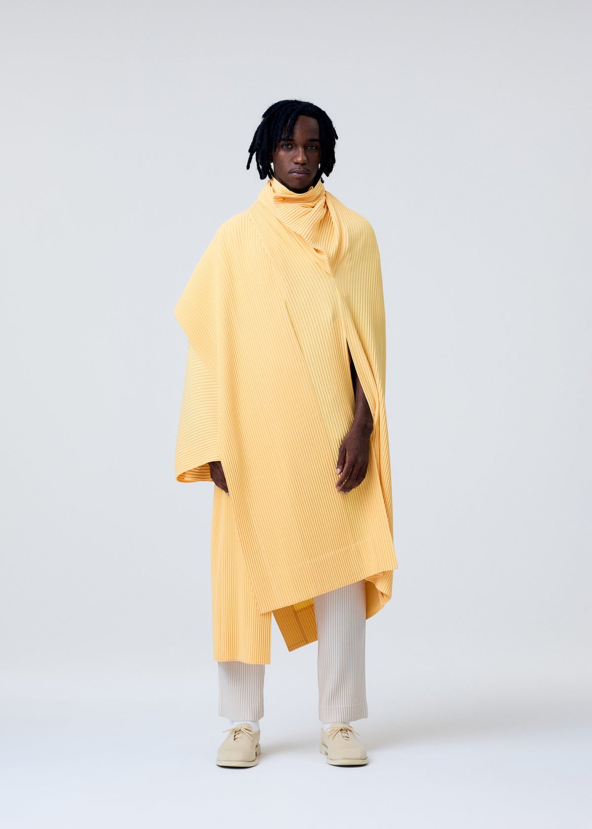A person with medium-length dreadlocks wears the HOMME PLISSÉ ISSEY MIYAKE WEAR OR WRAP—a flowing, pleated yellow poncho—over a matching stole wrap, cream pants, and beige shoes, standing against a plain light background. view 5 of 7