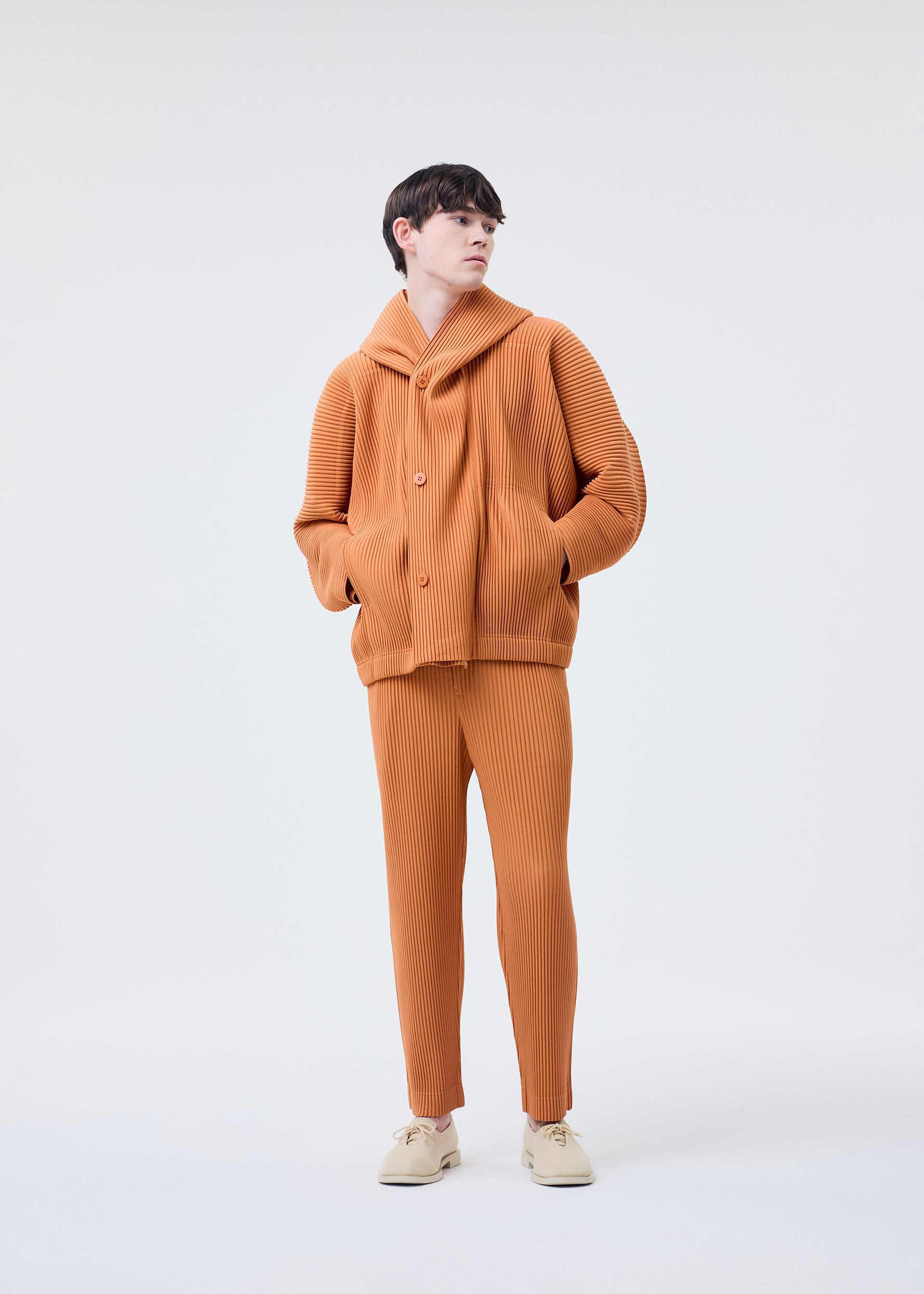 Zoomed view A person stands against a plain light background wearing the HOMME PLISSÉ ISSEY MIYAKE MONTHLY COLORS : MARCH matching ribbed quick-drying polyester orange jacket and pants set with cream shoes, looking slightly to the side with hands in pockets.
