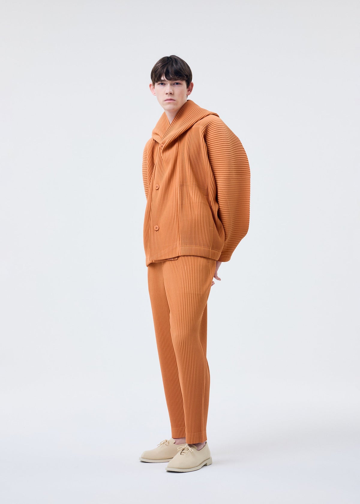 A person stands against a plain background, wearing HOMME PLISSÉ ISSEY MIYAKE’s MONTHLY COLORS : MARCH—an orange, textured polyester outfit with wide sleeves and matching pants, styled with light-colored shoes. view 7 of 7