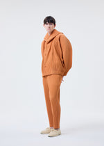 A person stands against a plain background, wearing HOMME PLISSÉ ISSEY MIYAKE’s MONTHLY COLORS : MARCH—an orange, textured polyester outfit with wide sleeves and matching pants, styled with light-colored shoes.