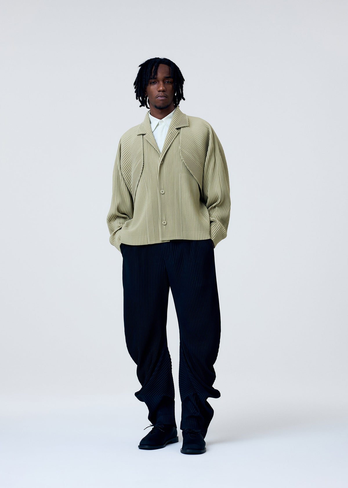 Wearing the HOMME PLISSÉ ISSEY MIYAKE COVER AND FOLD jacket with 3D pleats over a white shirt, loose black pleated pants, and black shoes, a person with short dreadlocks stands against a plain background. view 6 of 9