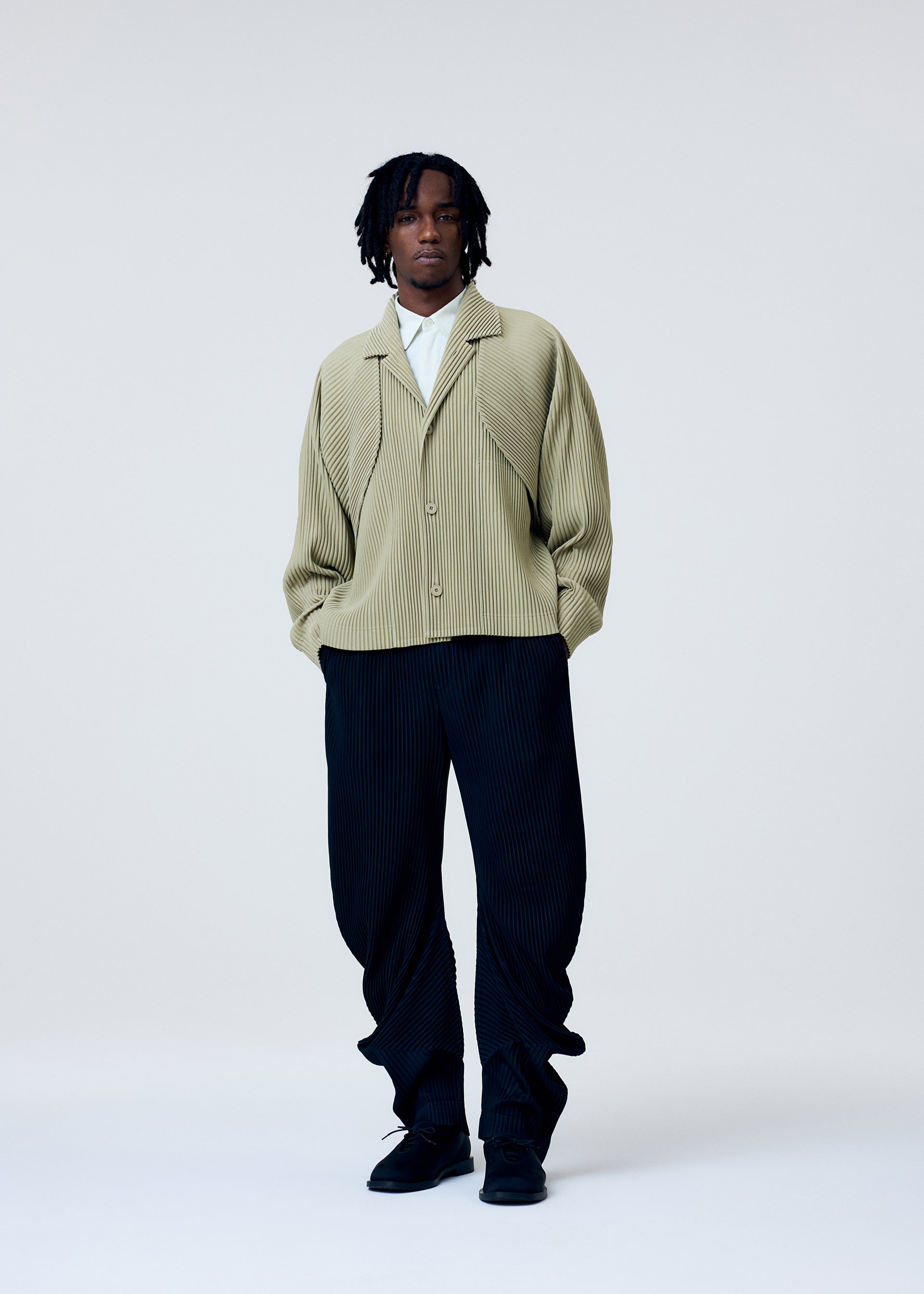 Zoomed view Wearing the HOMME PLISSÉ ISSEY MIYAKE COVER AND FOLD jacket with 3D pleats over a white shirt, loose black pleated pants, and black shoes, a person with short dreadlocks stands against a plain background.