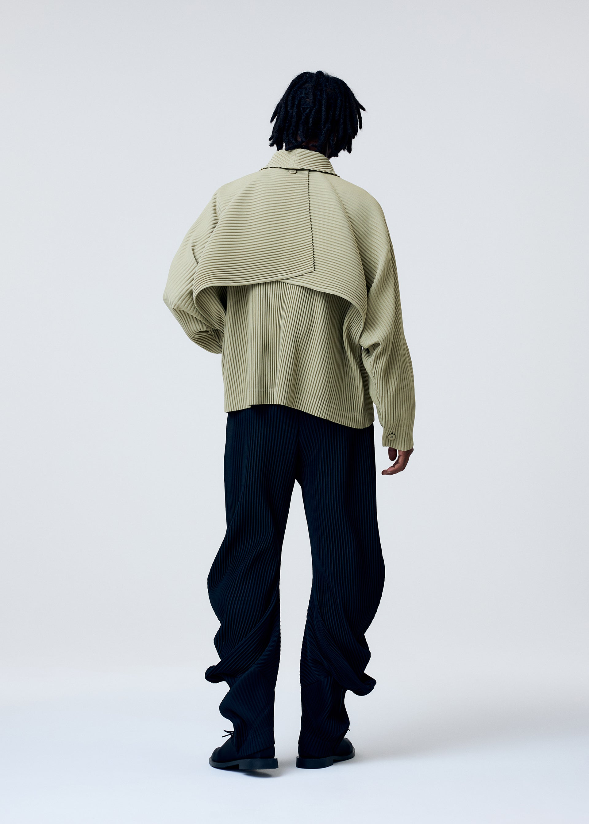 Zoomed view A person with short dreadlocks faces away, wearing the HOMME PLISSÉ ISSEY MIYAKE COVER AND FOLD jacket with 3D pleating over black pinstripe wide-leg pants, set against a plain light background.