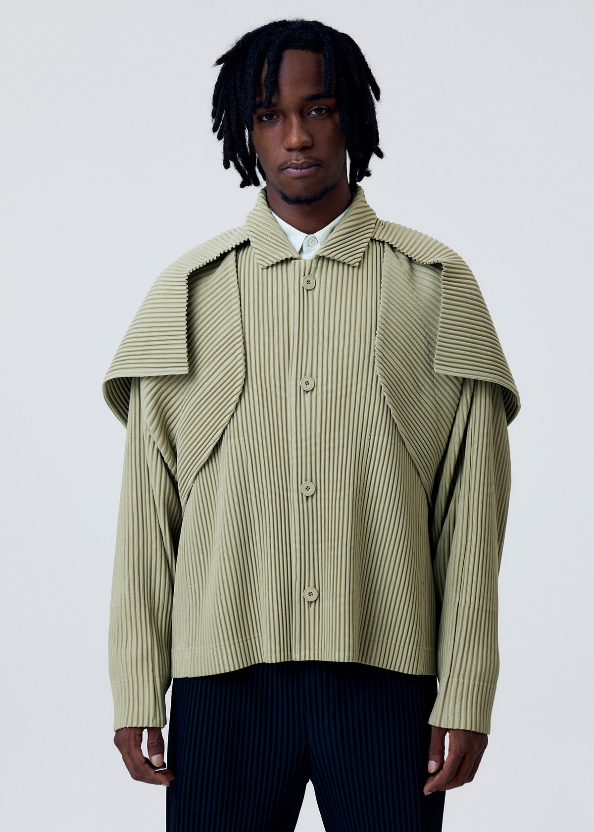 A man with medium-length dreadlocks models the HOMME PLISSÉ ISSEY MIYAKE COVER AND FOLD shirt—light olive green with 3D pleating and a layered shoulder—paired with dark striped pants, standing against a plain light background. view 8 of 9